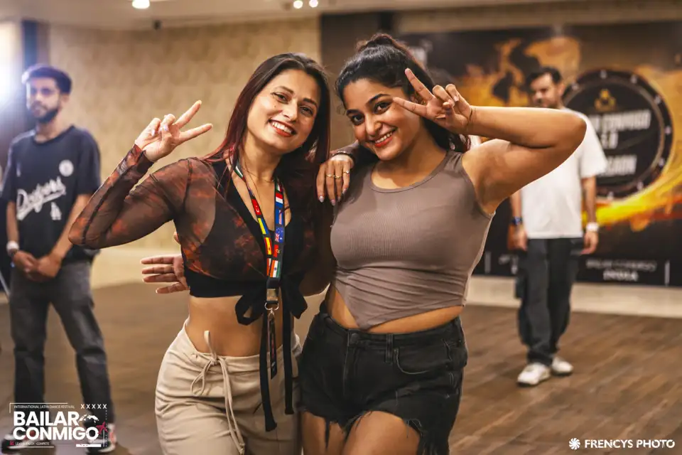 Two dancers arm in arm, laughing at the camera, with other people still on the floor behind them at a festival.