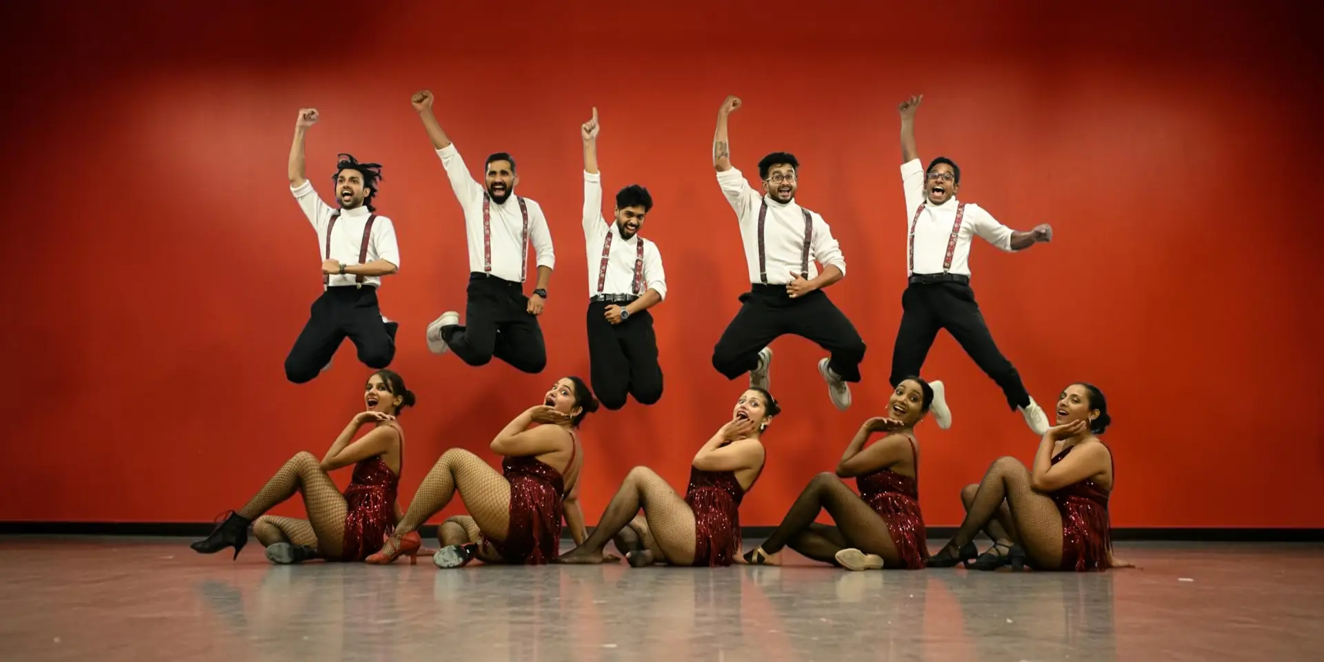 Ten dancers in matching red and black outfits — five leaping mid-air with arms raised, five posed on the floor in front of them — against a red studio wall.